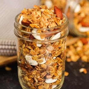 A glass mason jar filled with homemade pumpkin granola recipe, featuring oats, sliced almonds, and coconut flakes. Some granola is scattered on the dark surface around the jar.