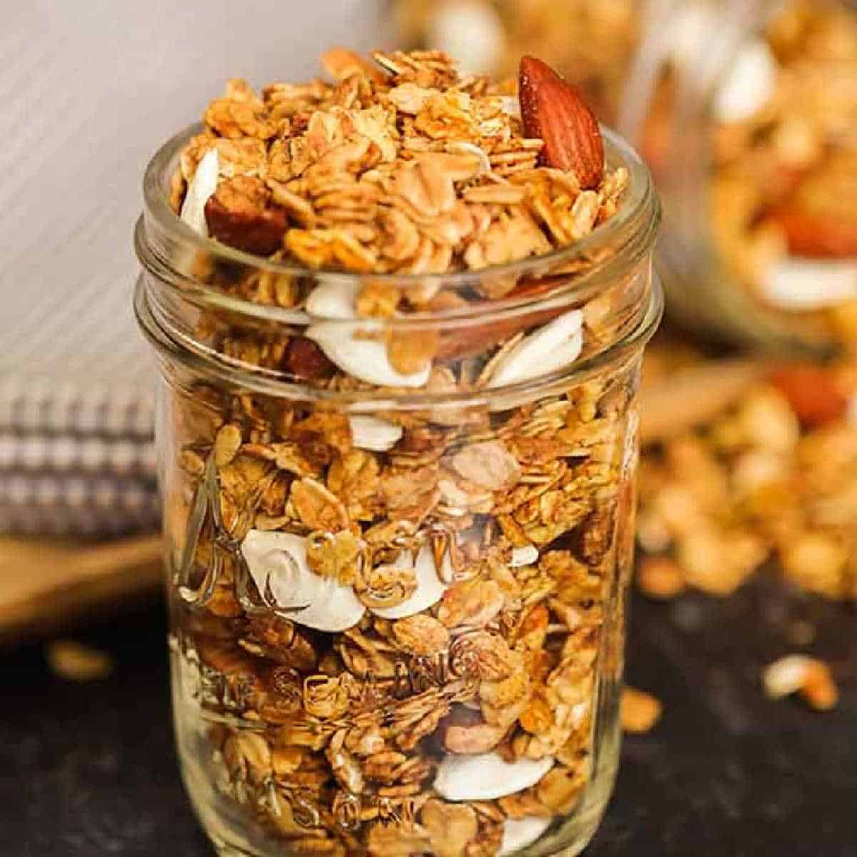 A glass mason jar filled with homemade pumpkin granola recipe, featuring oats, sliced almonds, and coconut flakes. Some granola is scattered on the dark surface around the jar.