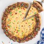 A large chocolate chip cookie cake, made using how to make cookie cake with cake mix, is decorated with chocolate frosting around the edge and topped with colorful candy sprinkles. One slice is being lifted from the cake.
