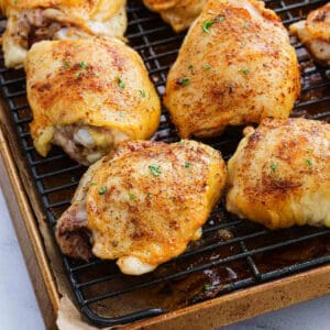 Several seasoned, oven baked chicken thighs with golden crispy skin are resting on a wire rack over a baking tray, garnished with a sprinkle of herbs.