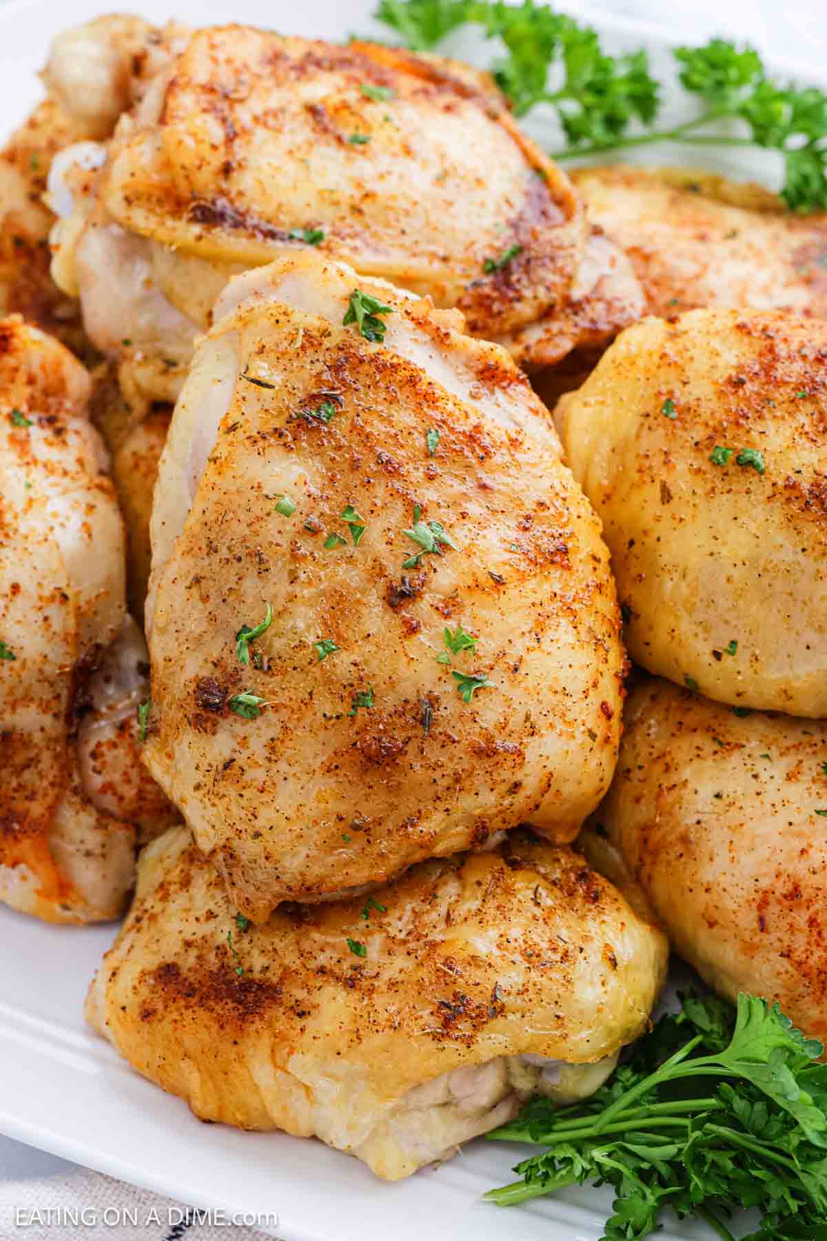 A close-up of several Oven Baked Chicken Thighs stacked on a white plate, seasoned to perfection and garnished with chopped parsley and fresh parsley sprigs.