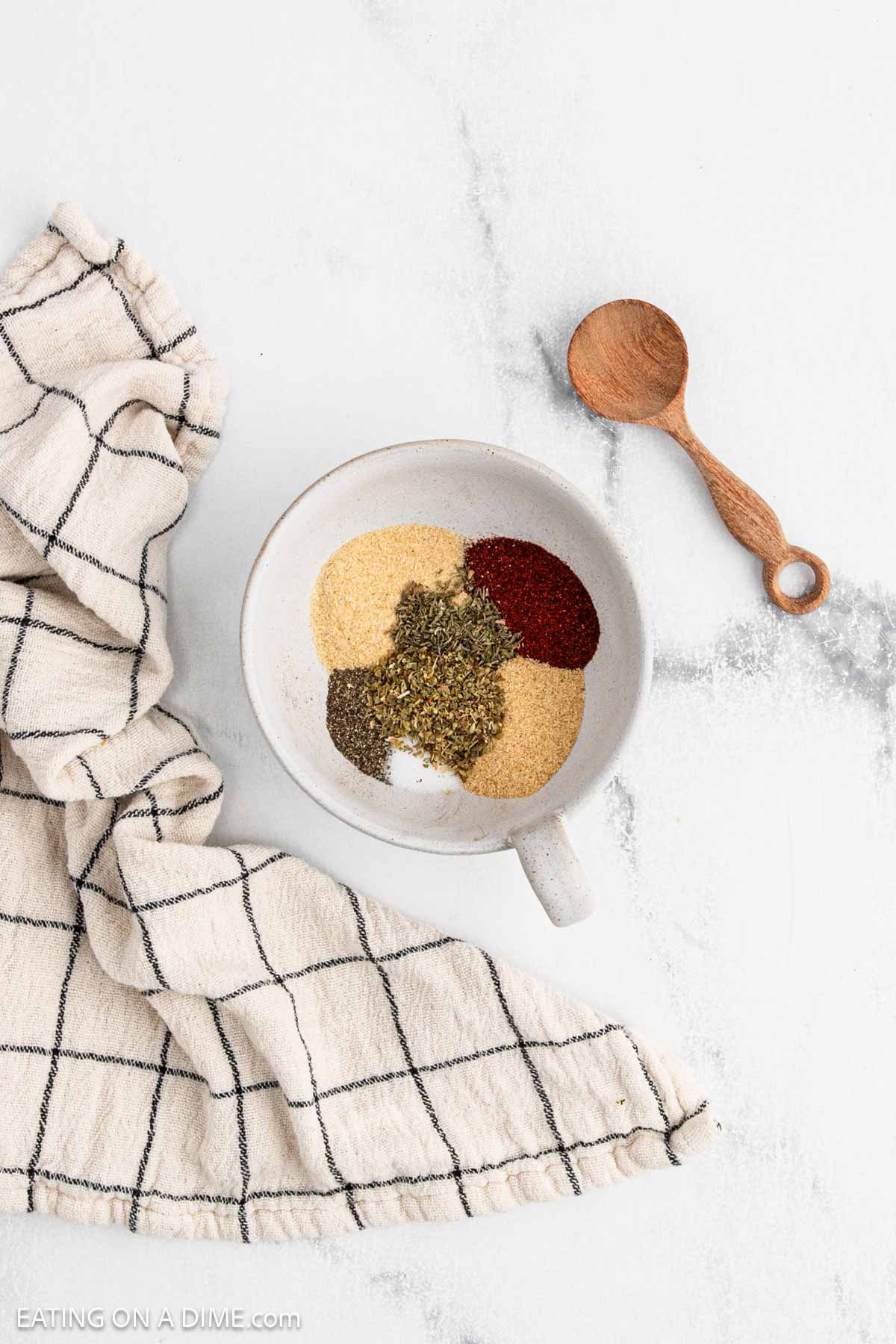 A white bowl containing separate piles of dried spices—perfect for seasoning Oven Baked Chicken Thighs—sits on a marble surface beside a checkered cloth and a small wooden spoon.