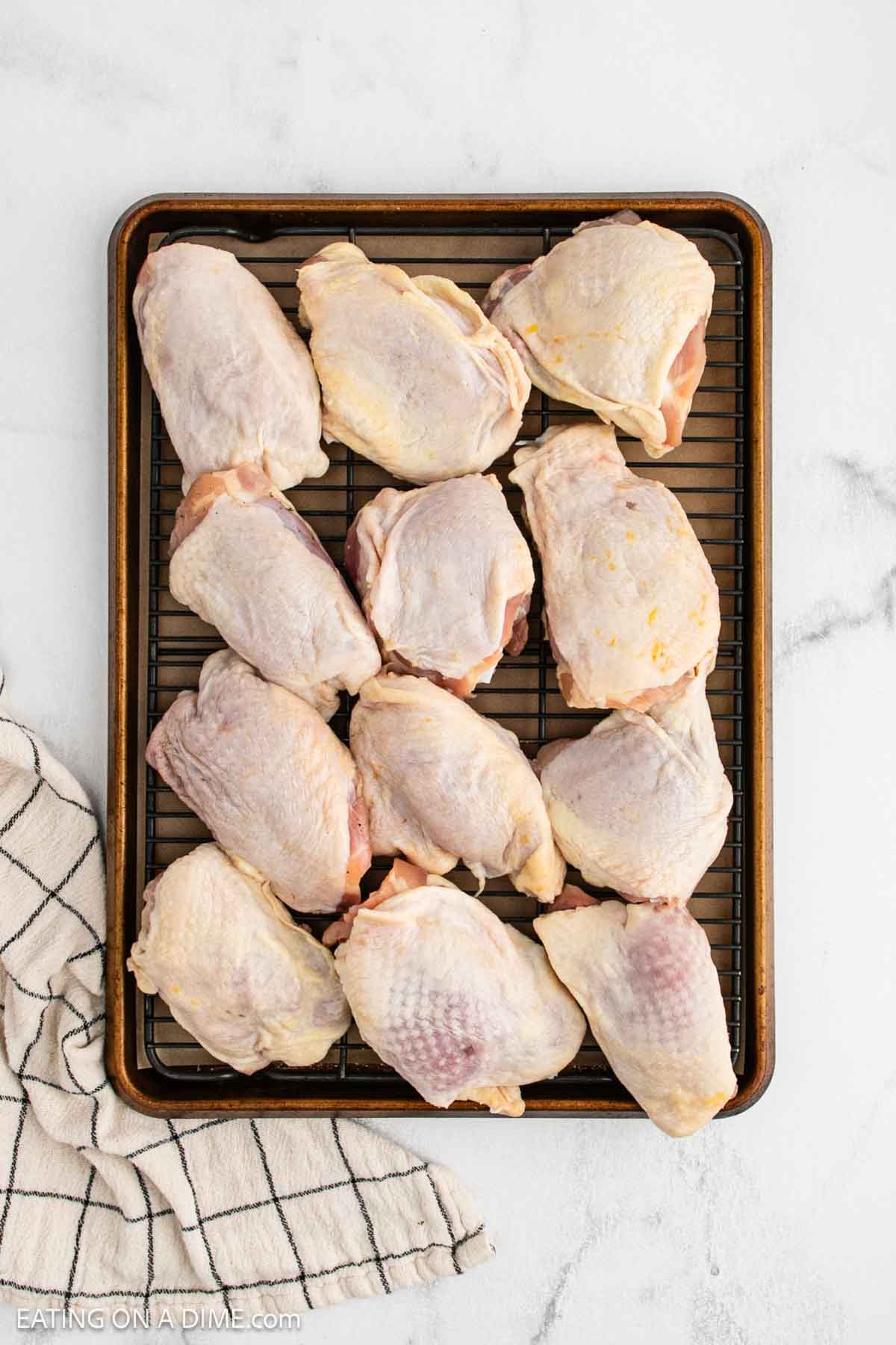 A baking tray with a wire rack holds twelve raw, skin-on chicken thighs, ready to become delicious Oven Baked Chicken Thighs. The tray sits on a white marble surface beside a folded striped kitchen towel.