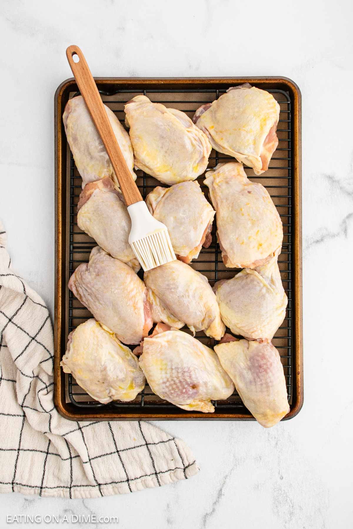 A baking sheet with a wire rack holds raw, seasoned Oven Baked Chicken Thighs. A pastry brush with a wooden handle rests on top, while a white kitchen towel with black stripes sits beside the tray on a white surface.