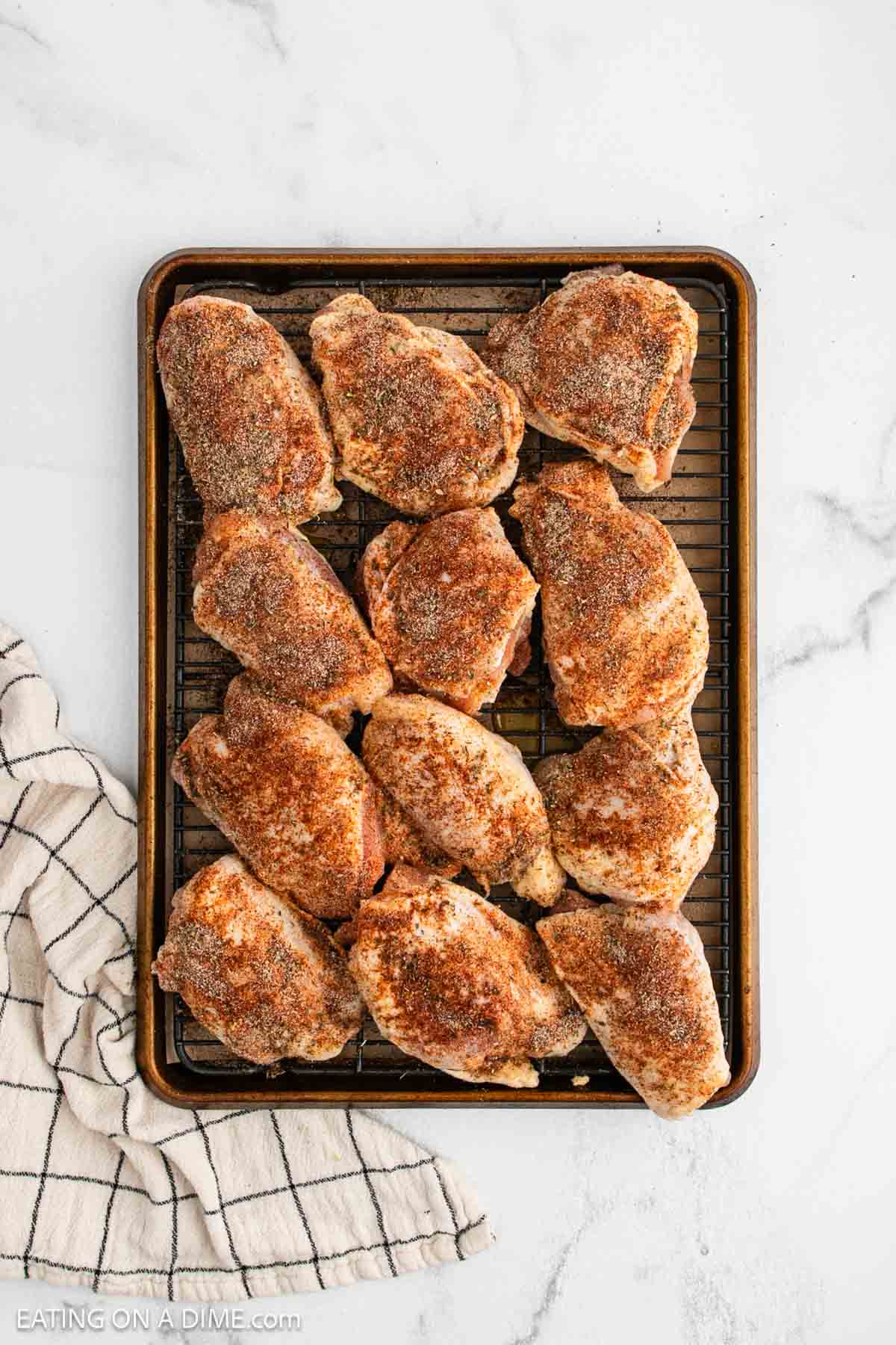 A baking sheet with a wire rack holds twelve seasoned, uncooked chicken thighs arranged in rows for oven baked chicken thighs. A white and black striped kitchen towel sits to the left on a white marble surface.