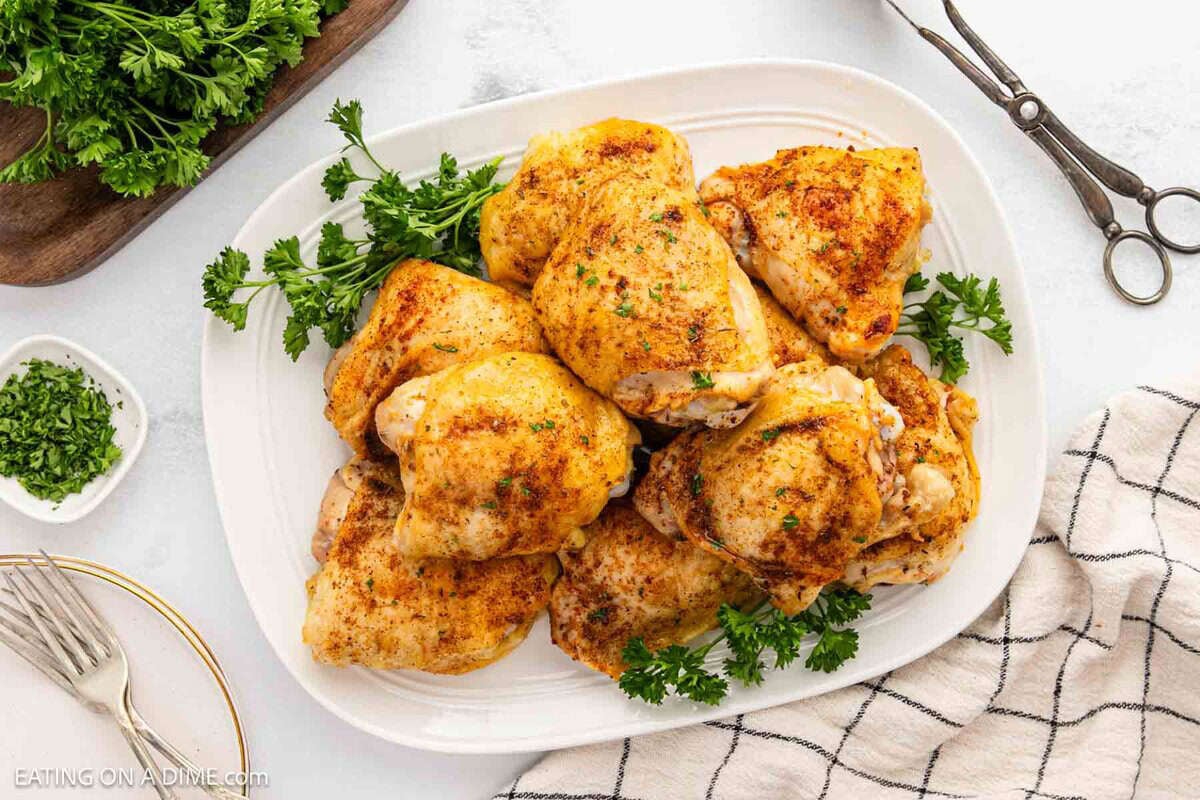 A white platter filled with Oven Baked Chicken Thighs, seasoned and garnished with fresh parsley, sits on a light surface beside a black-and-white checkered napkin, scissors, and a bunch of parsley.