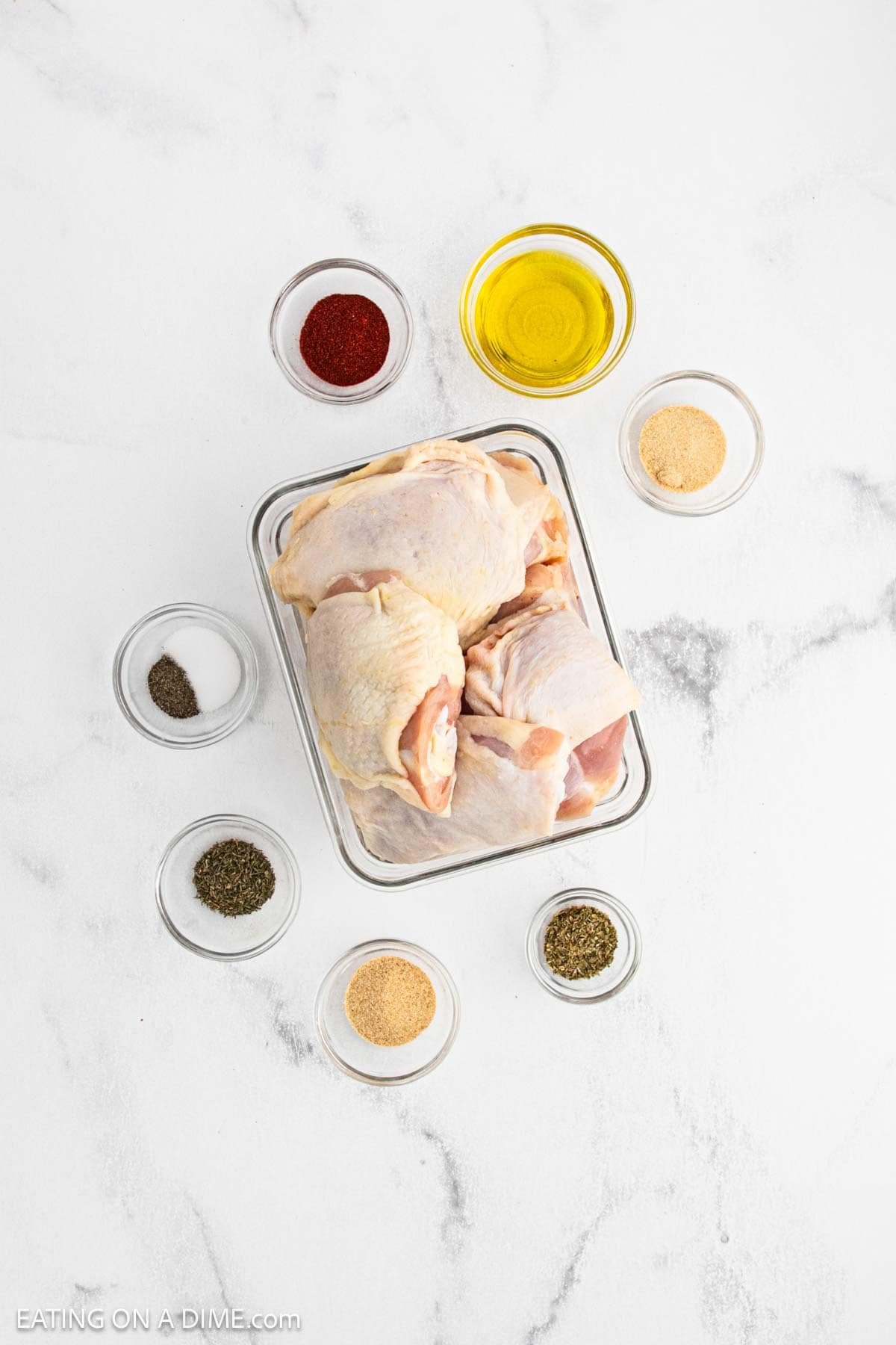 A glass container with raw chicken thighs for Oven Baked Chicken Thighs is surrounded by small bowls of olive oil, paprika, garlic powder, onion powder, dried basil, oregano, salt, and pepper on a white marble surface.