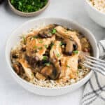 A bowl of Crock Pot Smothered Chicken over rice, topped with mushrooms and creamy sauce, garnished with chopped parsley. A fork rests on the bowl, with a napkin nearby and a dish of chopped herbs in the background.