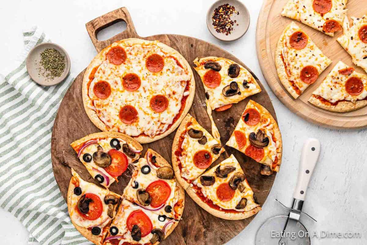 A variety of sliced Pita Bread Pizza with pepperoni, mushrooms, and olives are arranged on wooden boards. A pizza cutter, seasoning bowls, and a striped napkin are nearby on a white surface.