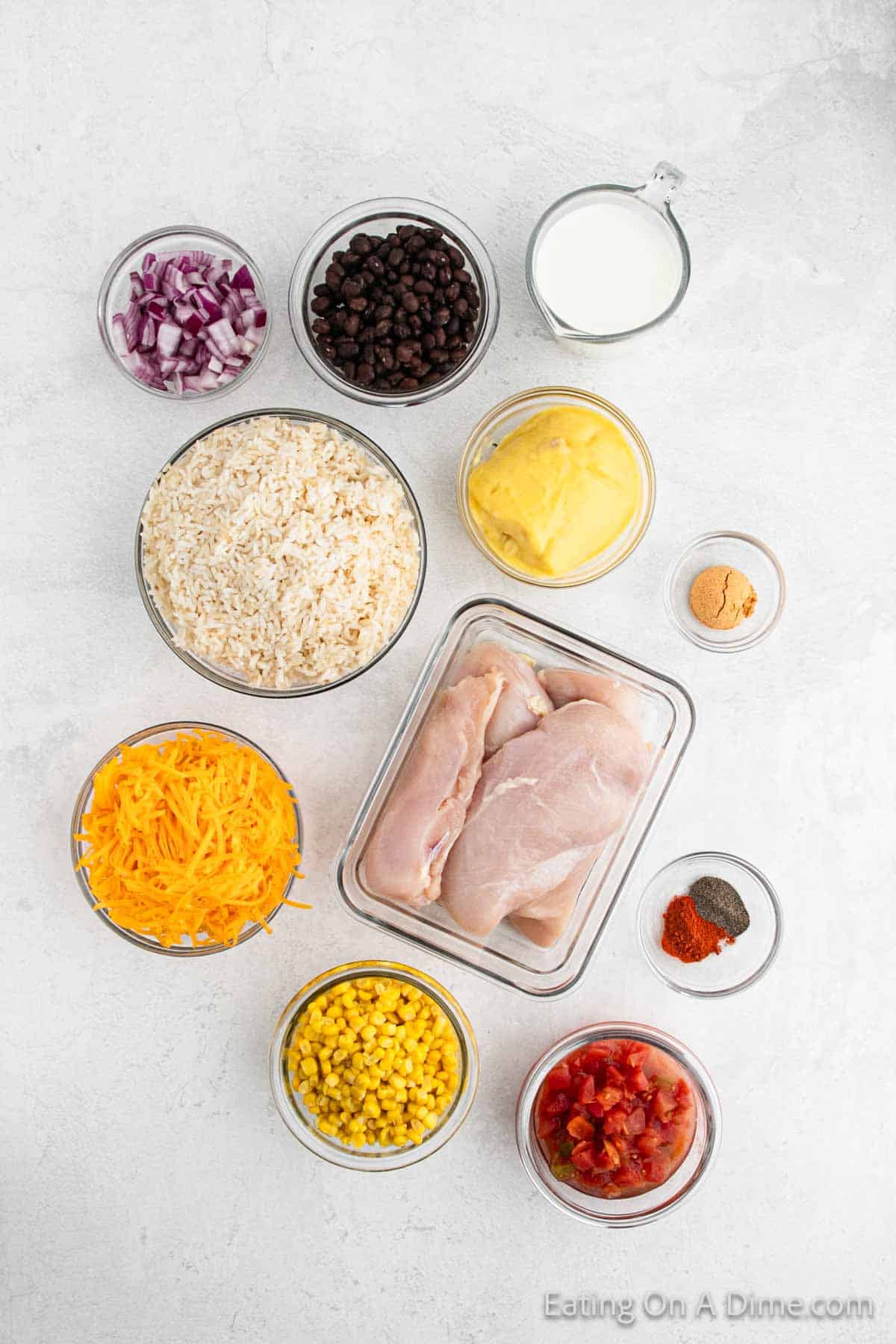 An overhead view of various ingredients on a white surface, perfect for making Southwest crock pot chicken and rice: raw chicken breasts, shredded cheddar cheese, cooked rice, corn, black beans, chopped red onions, diced tomatoes, a bowl of yellow cheese sauce, a small bowl of milk, and spices.