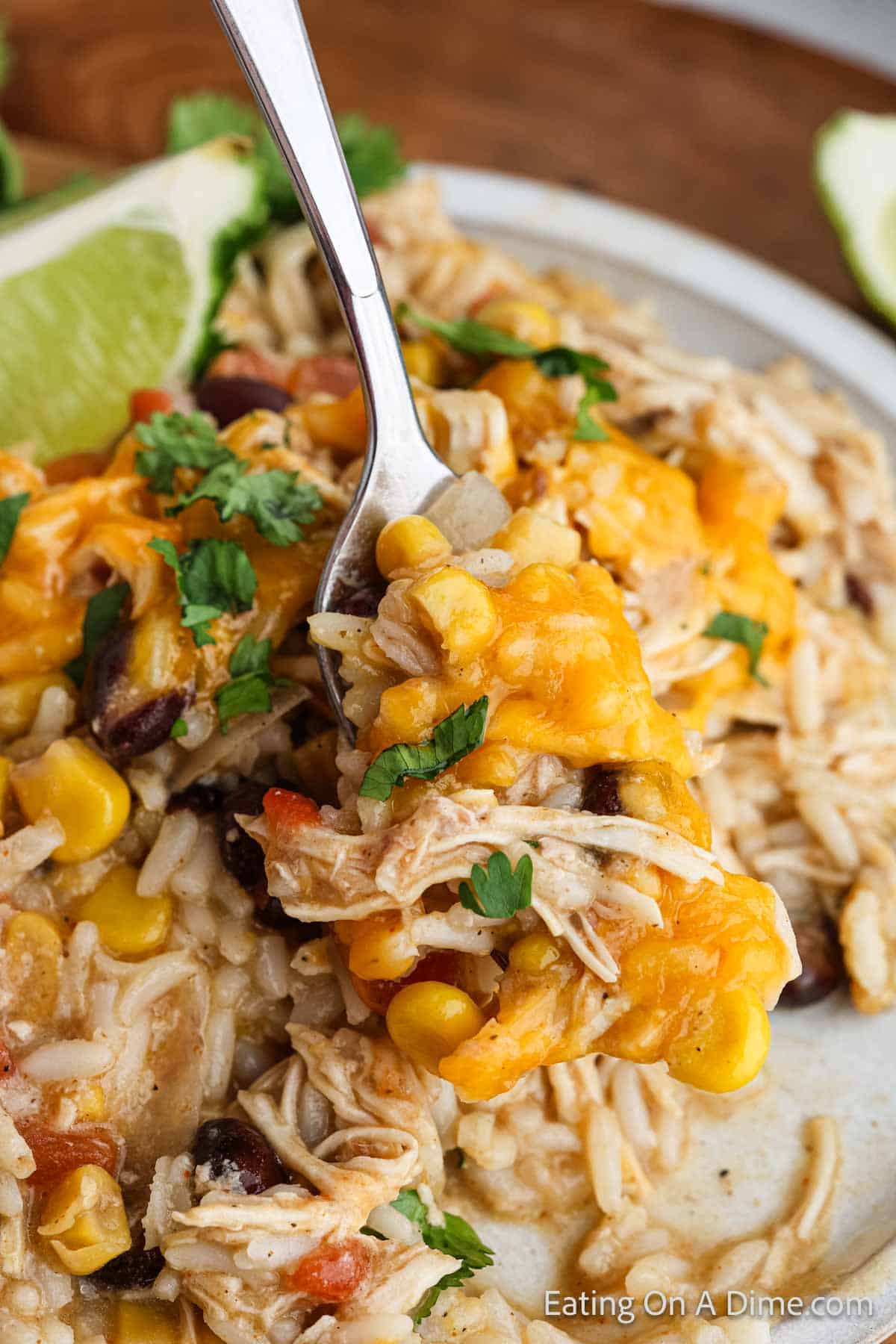 A close-up of a plate of cheesy Southwest crock pot chicken and rice casserole being scooped with a fork. The dish contains shredded chicken, melted cheese, corn, beans, and visible pieces of tomato, garnished with fresh cilantro and accompanied by lime wedges.