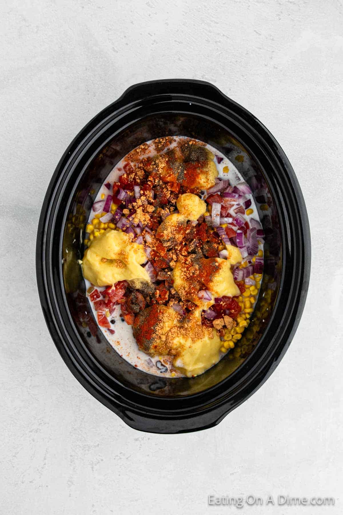 A black slow cooker filled with various ingredients, including red onions, corn, diced tomatoes, shredded cheese, cream, and assorted spices. The colorful mixture of Southwest crock pot chicken and rice is ready to be cooked on a light gray surface.