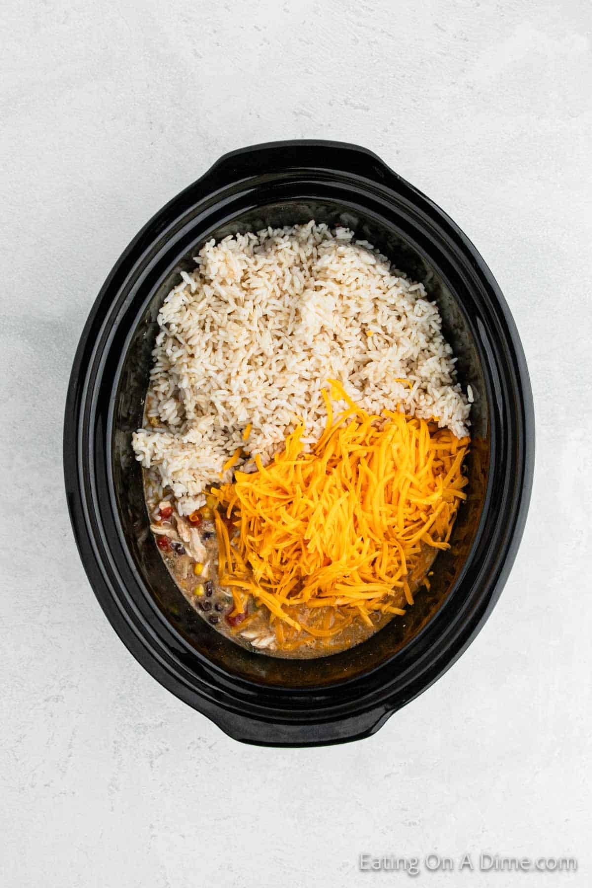 A slow cooker filled with a hearty mixture of cooked rice, shredded cheddar cheese, and tender Southwest crock pot chicken sits on a light-colored surface. The dish also includes vibrant pieces of vegetables and flavorful meat, perfect for a warm stew or comforting casserole.