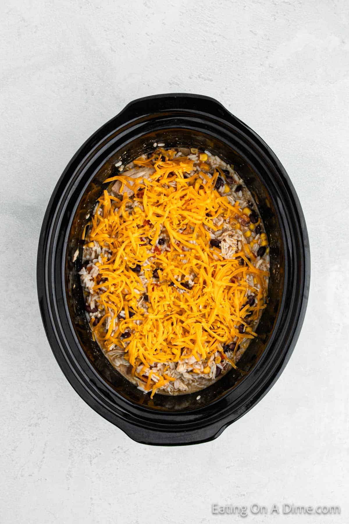 A black slow cooker filled with a Southwest crock pot chicken and rice mixture, including black beans, corn, and topped with shredded cheddar cheese. The background is a light grey speckled surface.