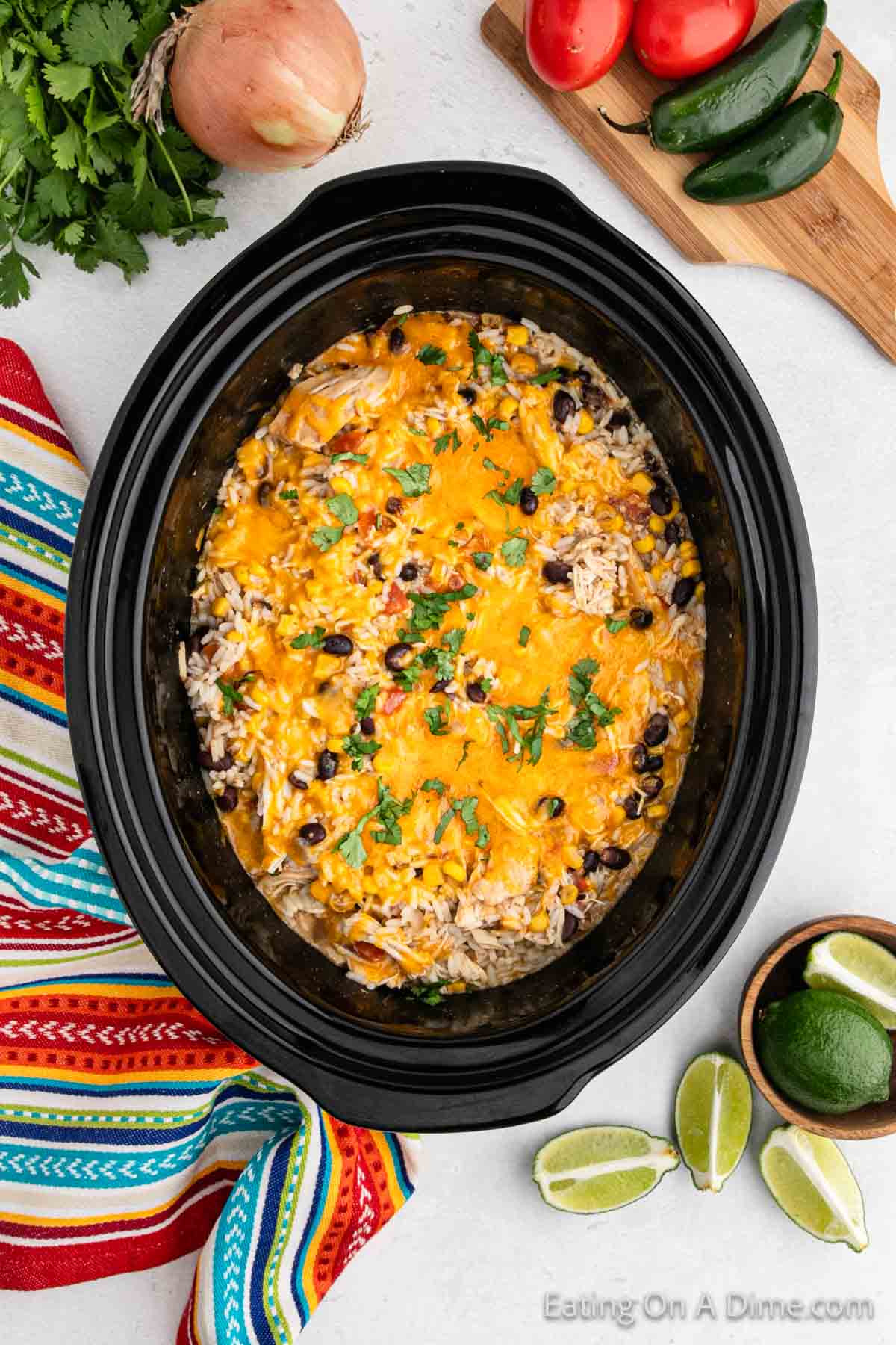A black slow cooker filled with Southwest crock pot chicken and rice, featuring a cheesy Mexican dish topped with melted cheddar cheese, rice, black beans, corn, and sprinkled with cilantro. Surrounding the slow cooker are cilantro, an onion, tomatoes, jalapeños, lime wedges, and a colorful striped cloth.