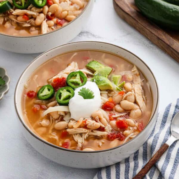A bowl of crock pot white chicken taco chili, topped with sliced jalapeños, diced avocado, shredded cheese, and a dollop of sour cream, with a sprig of cilantro. A striped napkin and jalapeño are nearby.