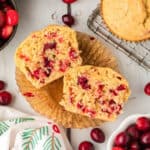 A cranberry muffin cut in half sits on an unwrapped muffin liner, revealing the moist interior packed with tart cranberries. Whole cranberries and extra cranberry muffins are scattered across a white surface nearby.