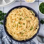 A bowl of Garlic Parmesan Pasta mixed with creamy white sauce, garnished with chopped parsley. Plates, a spoon, shredded cheese, and extra parsley are arranged around the bowl on a tiled surface.