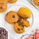 Three pumpkin chocolate chip muffins are on a white plate, one broken in half to reveal the melty chocolate chips inside. More pumpkin chocolate chip muffins, a bowl of chocolate chips, and a colorful napkin sit nearby on the marble surface.