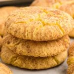 Three golden-brown snickerdoodle cake mix cookies are stacked on top of each other, showing a cracked, cinnamon-sugar coated surface. Other cookies from this easy recipe are visible in the blurred background.