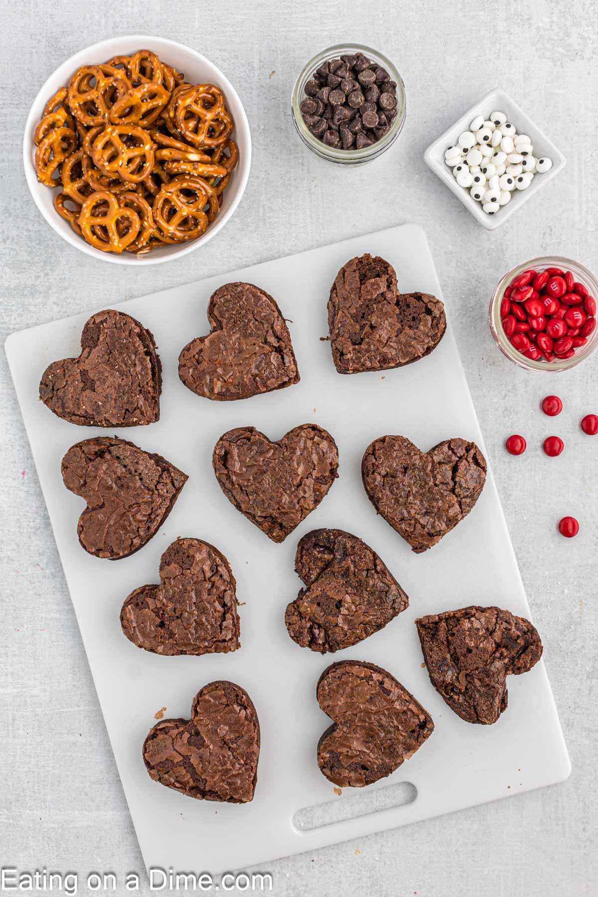 Heart shaped brownies on a cutting board with bowls of pretzels, chocolate chips, candy eyes and red M&M