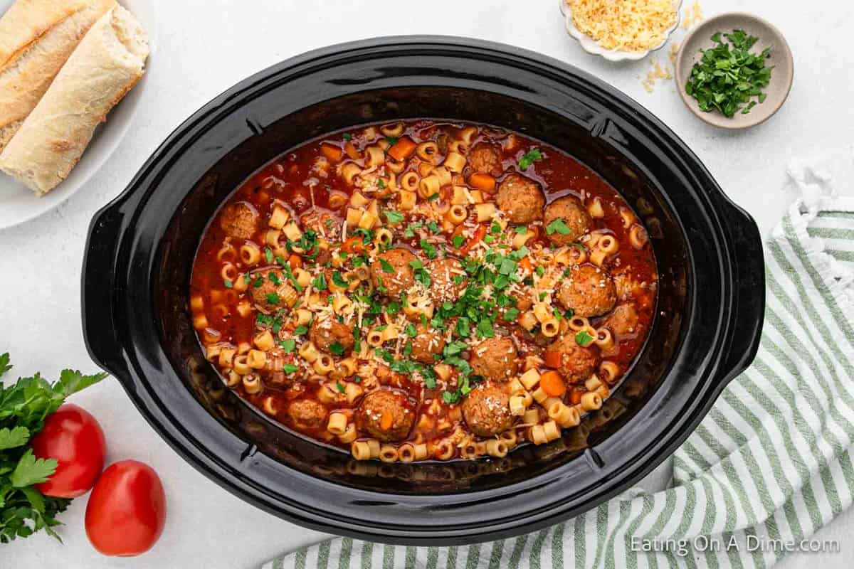 A Crock Pot brimming with Italian meatball pasta soup, garnished with chopped herbs, sits invitingly. Surrounding it are bread slices, grated cheese, and parsley on a pristine white surface. Nearby, a striped cloth adds an extra touch of cozy charm.
