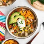 A bowl of Mexican chicken soup recipe, topped with avocado slices, sour cream, cilantro, and crispy tortilla strips, is surrounded by a colorful napkin, a spoon, and part of a cutting board with fresh cilantro.