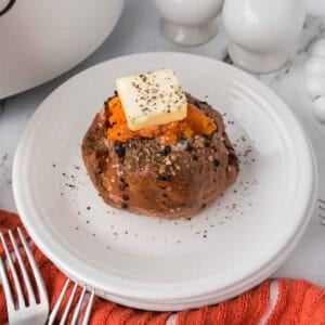 An Air Fryer Baked Sweet Potato topped with a pat of butter and black pepper sits on a white plate, next to orange napkins and silver forks. Salt and pepper shakers are in the background.