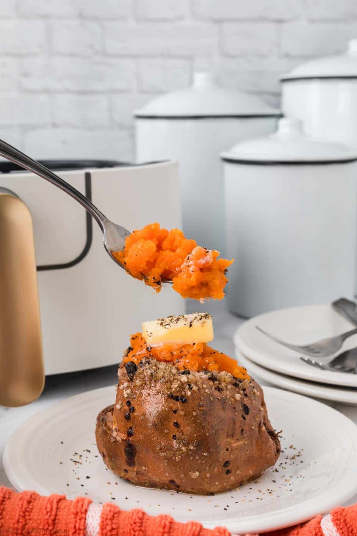 A fork holds a bite of mashed sweet potato above an Air Fryer Baked Sweet Potato topped with butter and black pepper, served on a white plate with a red towel and white canisters in the background.
