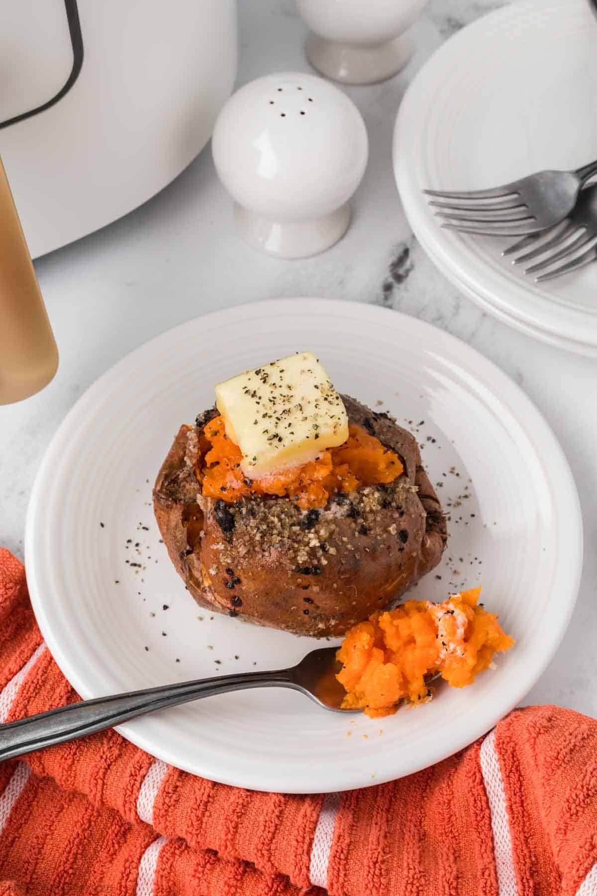 An Air Fryer Baked Sweet Potato sits on a white plate, topped with butter and black pepper, with a fork and orange napkin nearby. Salt and pepper shakers and a stack of plates with forks appear in the background.