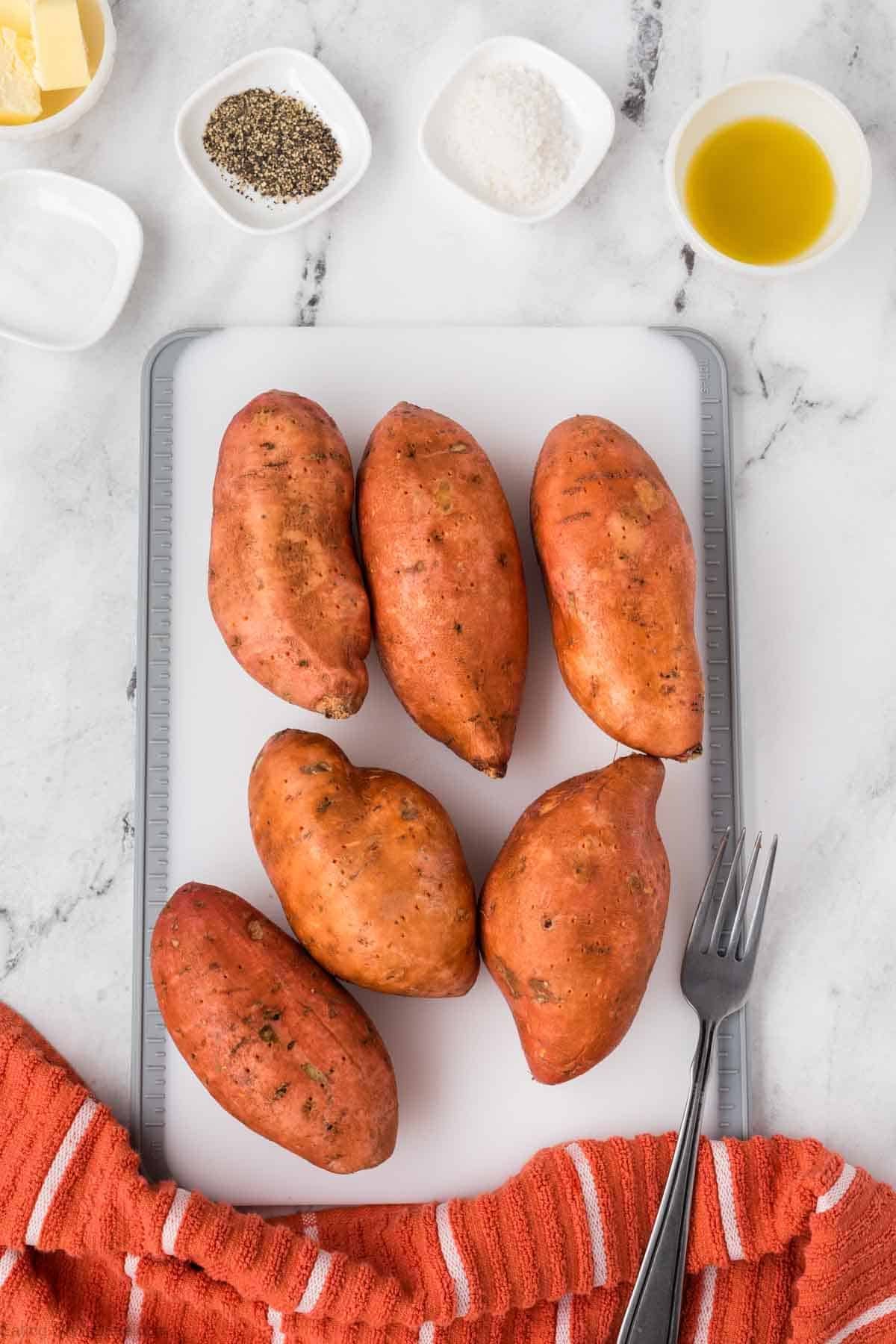 Six whole sweet potatoes are arranged on a white cutting board with a gray border, ready to become delicious Air Fryer Baked Sweet Potatoes. Nearby are bowls of olive oil, salt, pepper, and butter, plus a fork and an orange-striped towel on marble.