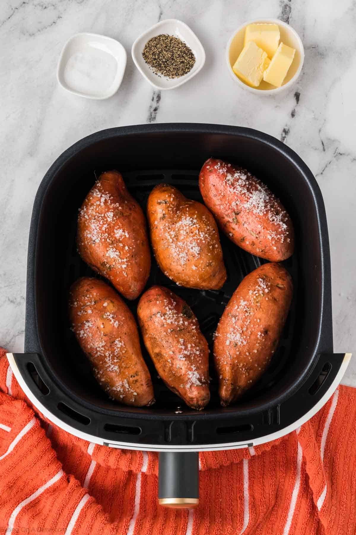 Six whole sweet potatoes sprinkled with salt are arranged in the basket, ready for Air Fryer Baked Sweet Potato perfection. Nearby, small bowls of butter, black pepper, and salt sit on a marble countertop with an orange kitchen towel.