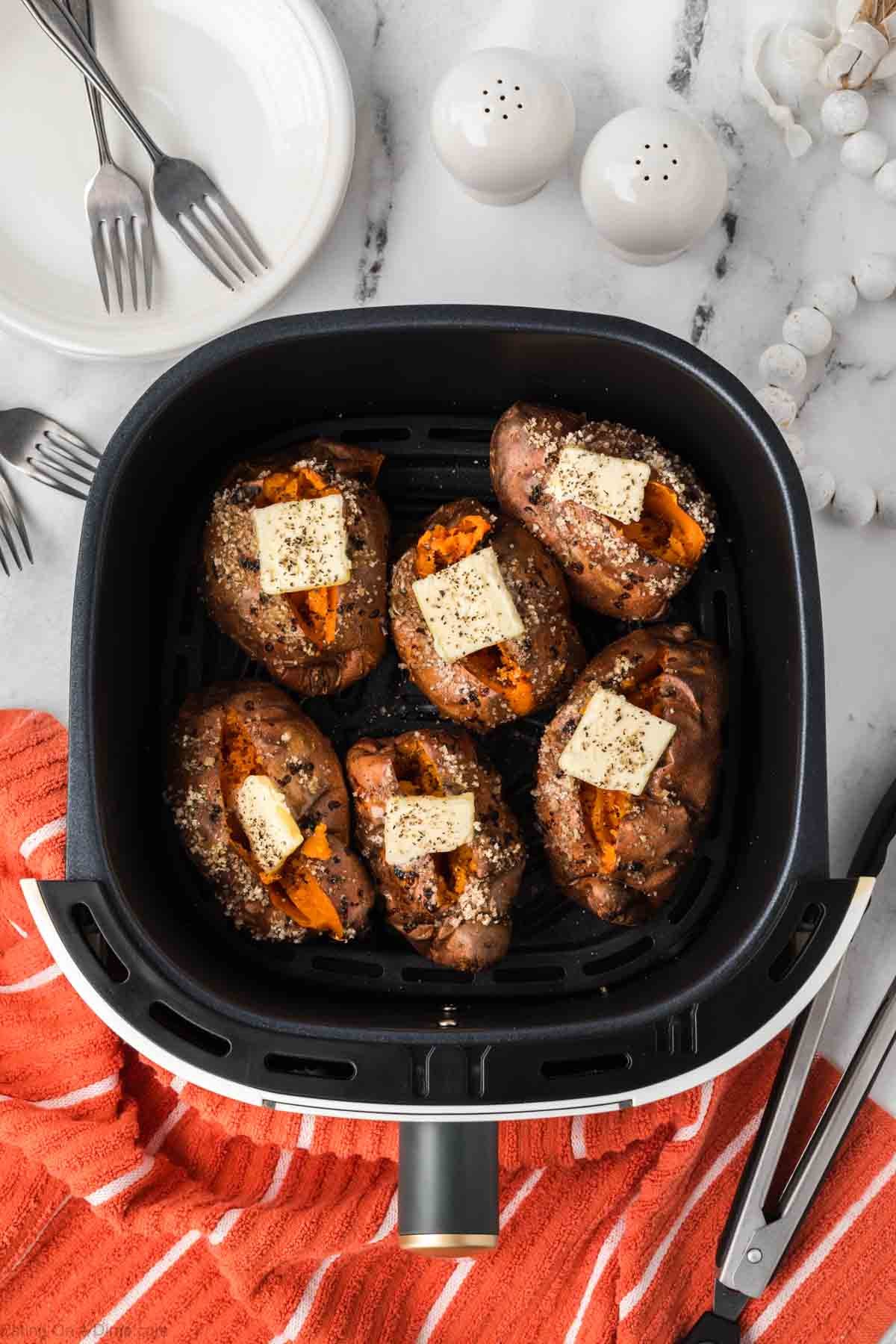 Five Air Fryer Baked Sweet Potatoes sit in the basket, each topped with a pat of butter and sprinkled herbs. Plates, forks, and salt and pepper shakers are arranged on the white countertop nearby.