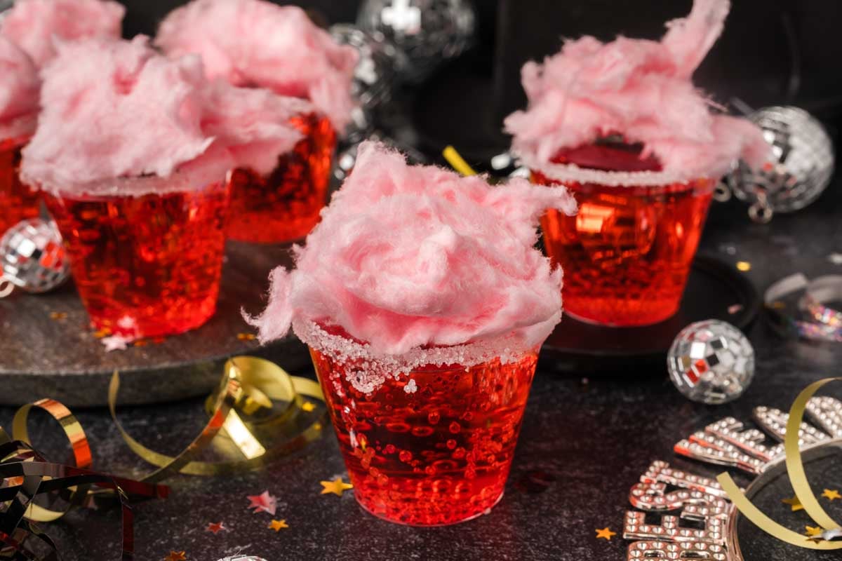 Clear plastic cups filled with a red sparkling cotton candy drink recipe, each topped with fluffy pink cotton candy. New Year's Eve decorations, party hats, streamers, and confetti surround the festive drinks on a dark surface.