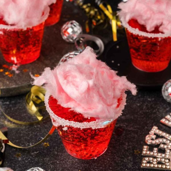 A clear plastic cup filled with red sparkling Cotton Candy Drink, topped with pink cotton candy and a sugar-coated rim, sits on a festive table adorned with gold ribbons and party decorations.