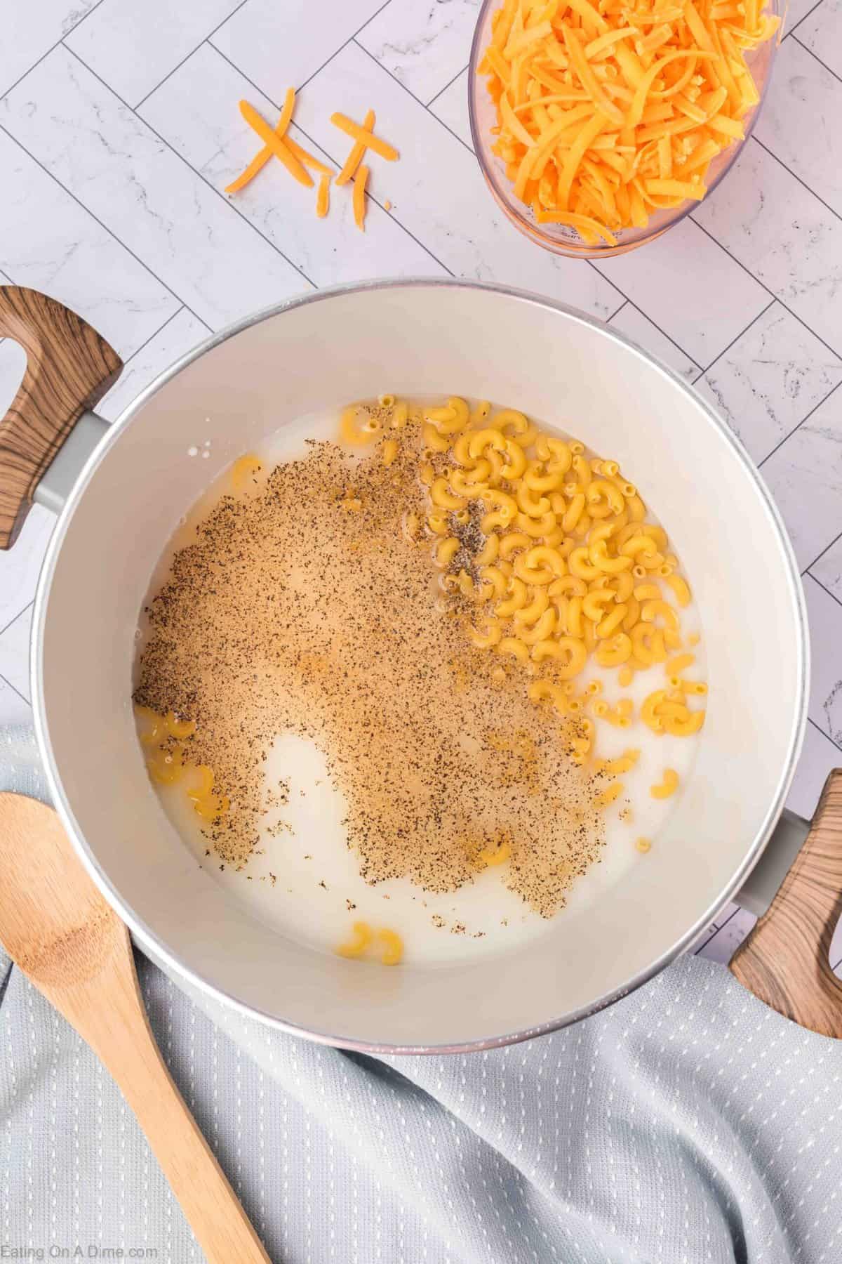 A pot on a white tiled countertop contains milk, elbow macaroni, and black pepper, ready to be cooked.
