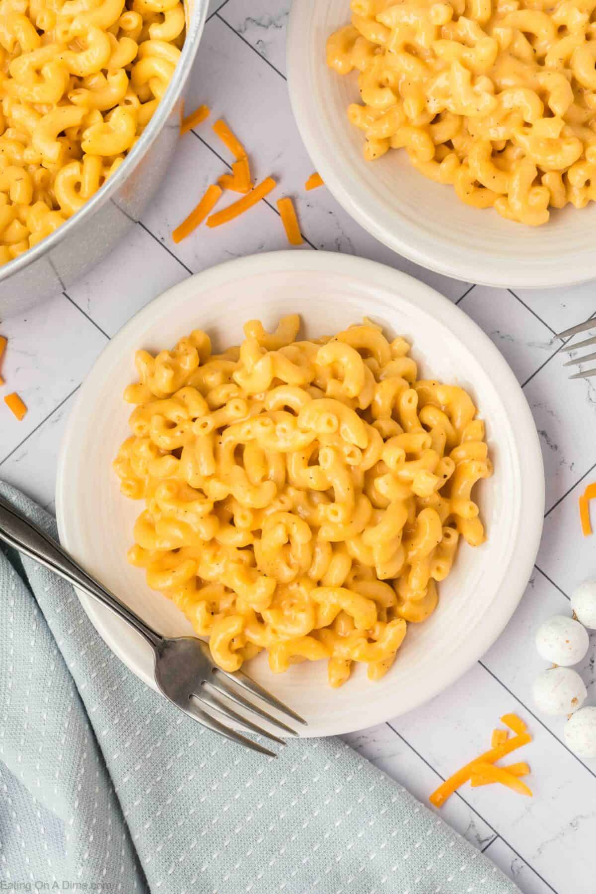 Overhead view of two plates and a pot filled with an easy homemade macaroni and cheese recipe. Forks are placed on the plates. The background features a blue cloth and scattered pieces of shredded cheddar cheese.