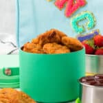 A green container filled with crispy chicken nuggets sits next to a small metal bowl of strawberries, a cup of dipping sauce, and a colorful backpack in the background—perfect inspiration if you’re wondering how to pack chicken nuggets for lunch.