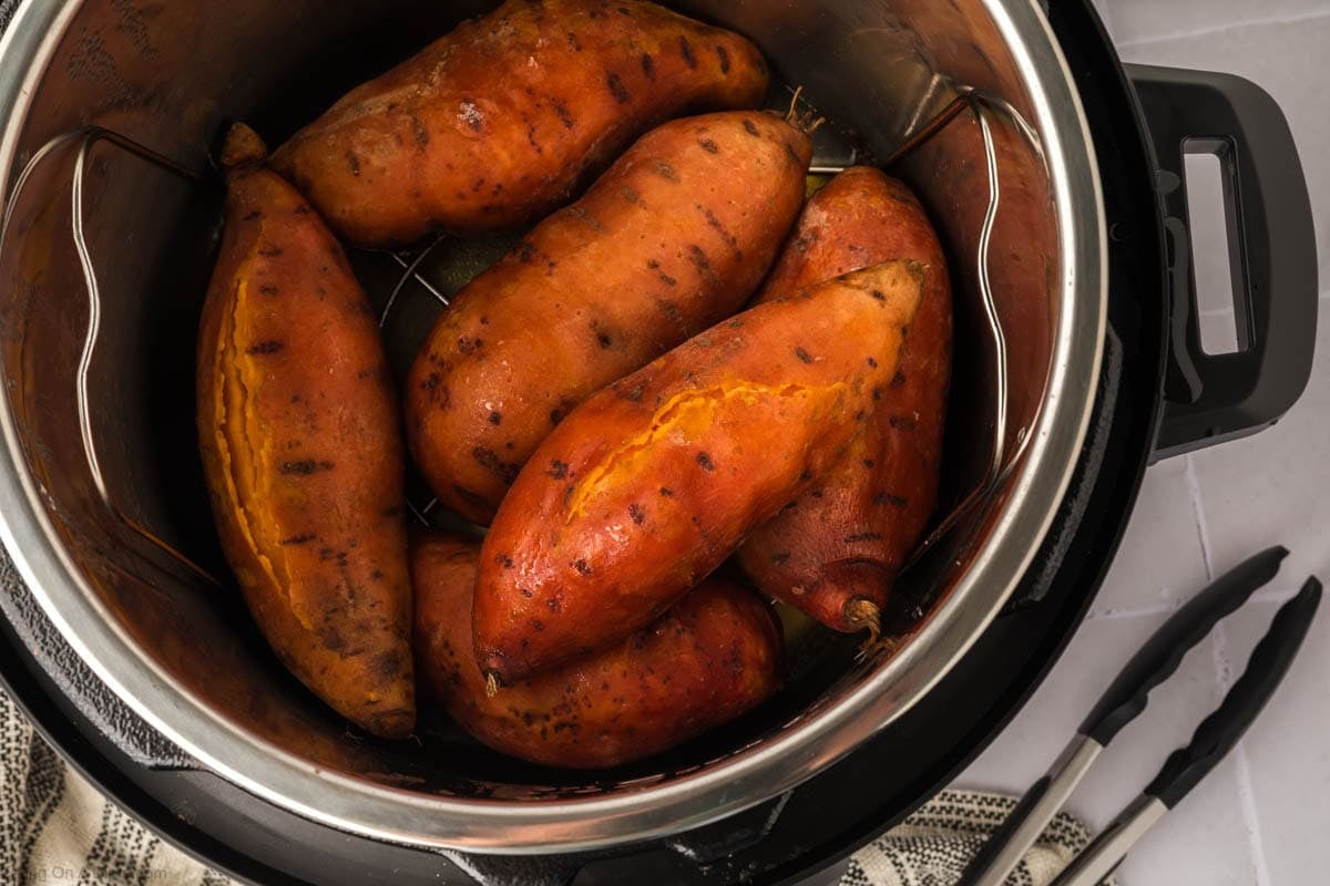 Six Instant Pot Sweet Potatoes with slightly cracked skins rest inside the appliance, while a pair of metal tongs and a kitchen towel sit nearby on the counter.