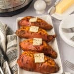 Four baked Instant Pot Sweet Potatoes on a white rectangular plate, each topped with butter and black pepper. Forks, a stick of butter, salt and pepper shakers, and an Instant Pot sit nearby on the table.