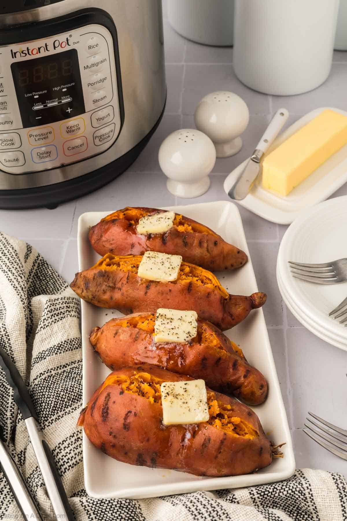 Four Instant Pot Sweet Potatoes topped with butter and black pepper on a white rectangular plate, next to an Instant Pot, a stick of butter, salt and pepper shakers, forks, and a striped kitchen towel.