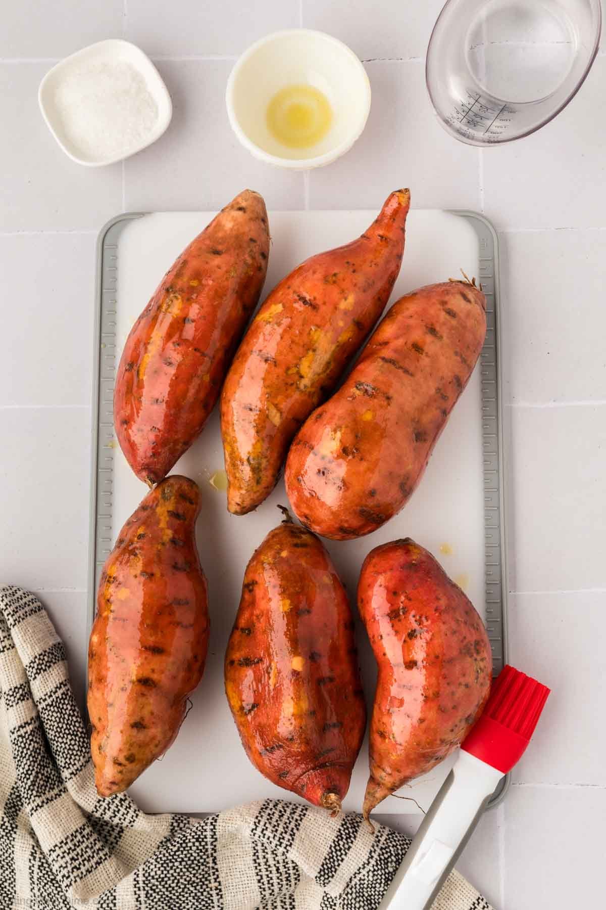 Six whole, shiny sweet potatoes sit on a white cutting board, ready to become Instant Pot Sweet Potatoes. They’re surrounded by a small bowl of oil, salt, water in a measuring cup, a pastry brush, and a striped kitchen towel on a light surface.