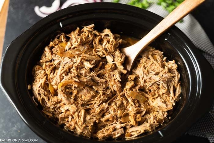 Shredded pork in a black crock pot with a wooden spoon in it 