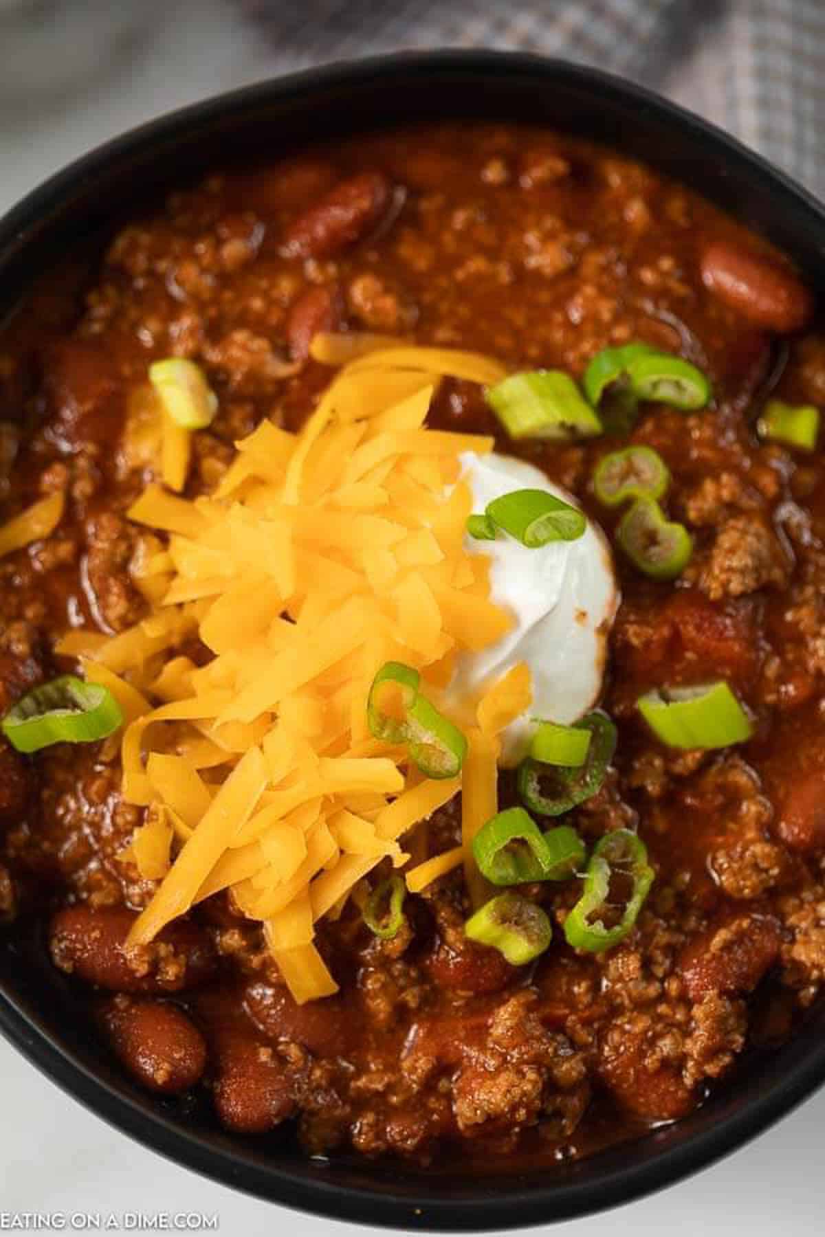 Close up image of chili in a black bowl topped with cheese, sour cream and green onions. 