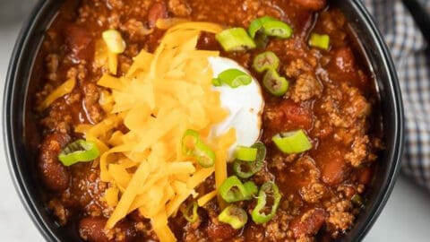 Close up image of chili in a black bowl topped with cheese, sour cream and green onions.