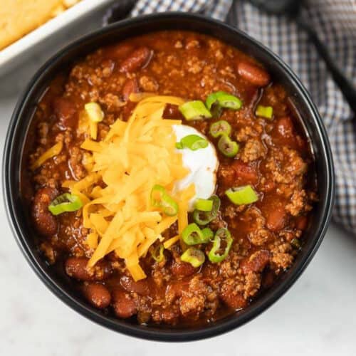 Close up image of chili in a black bowl topped with cheese, sour cream and green onions.