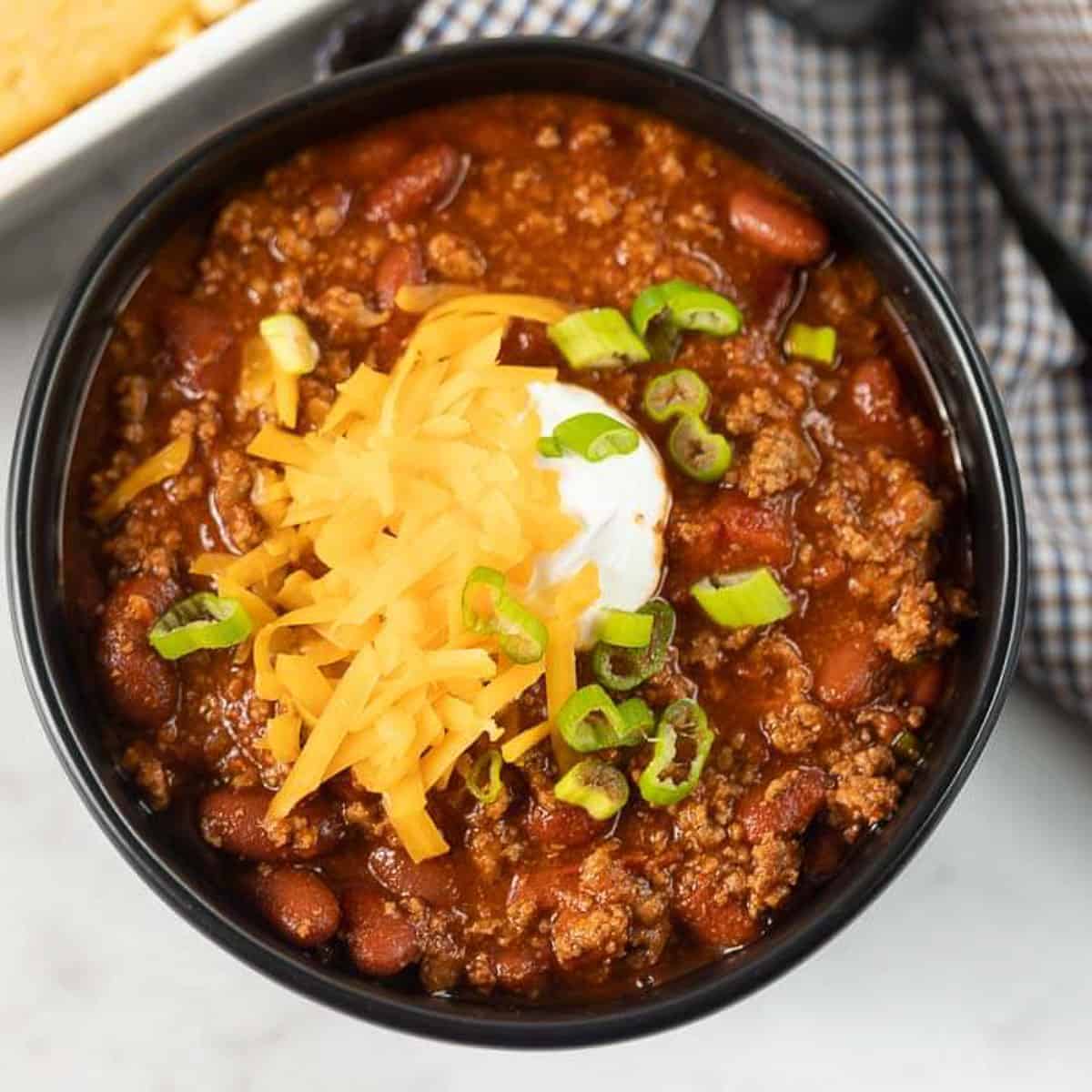 Close up image of chili in a black bowl topped with cheese, sour cream and green onions. 