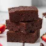 Three thick, fudgy low calorie brownies are stacked on a white surface, with fresh strawberry halves scattered around them.