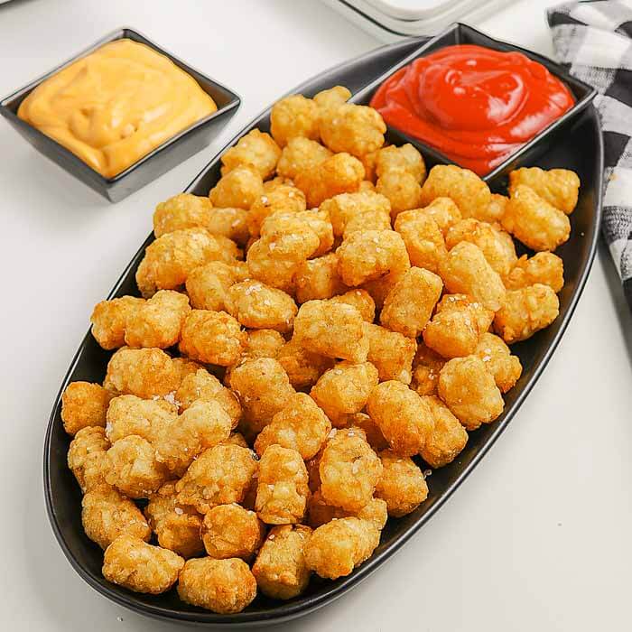Close up image of a platter of air fryer tator tots with a side of cheese sauce and ketchup. 