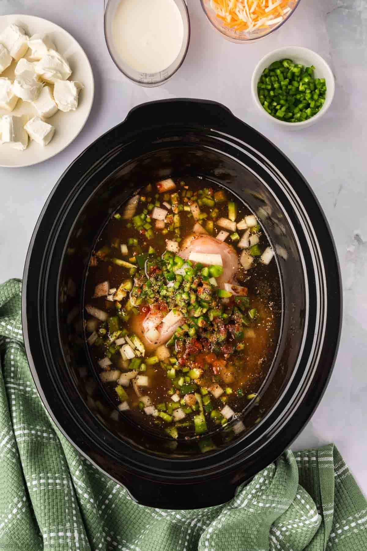 Chicken broth poured over the chicken, diced vegetables and seasoning in the crock pot with a plate of diced cream cheese, heavy cream, shredded cheese and diced jalapenos on the side